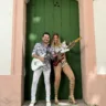 O músico Ximbinha posa em frente a uma porta antiga verde, segurando uma guitarra branca. Ao lado dele, Bia Villa-Chan, uma mulher de cabelos louros, segura uma guitarra vermelha.