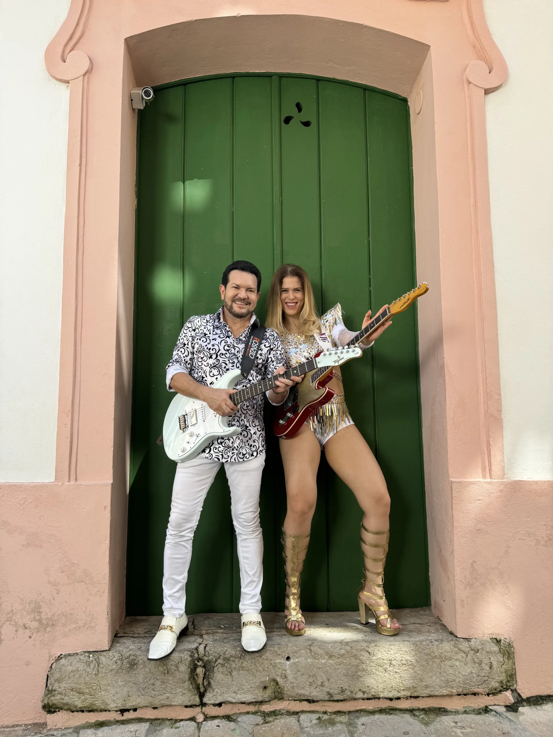 O músico Ximbinha posa em frente a uma porta antiga verde, segurando uma guitarra branca. Ao lado dele, Bia Villa-Chan, uma mulher de cabelos louros, segura uma guitarra vermelha.