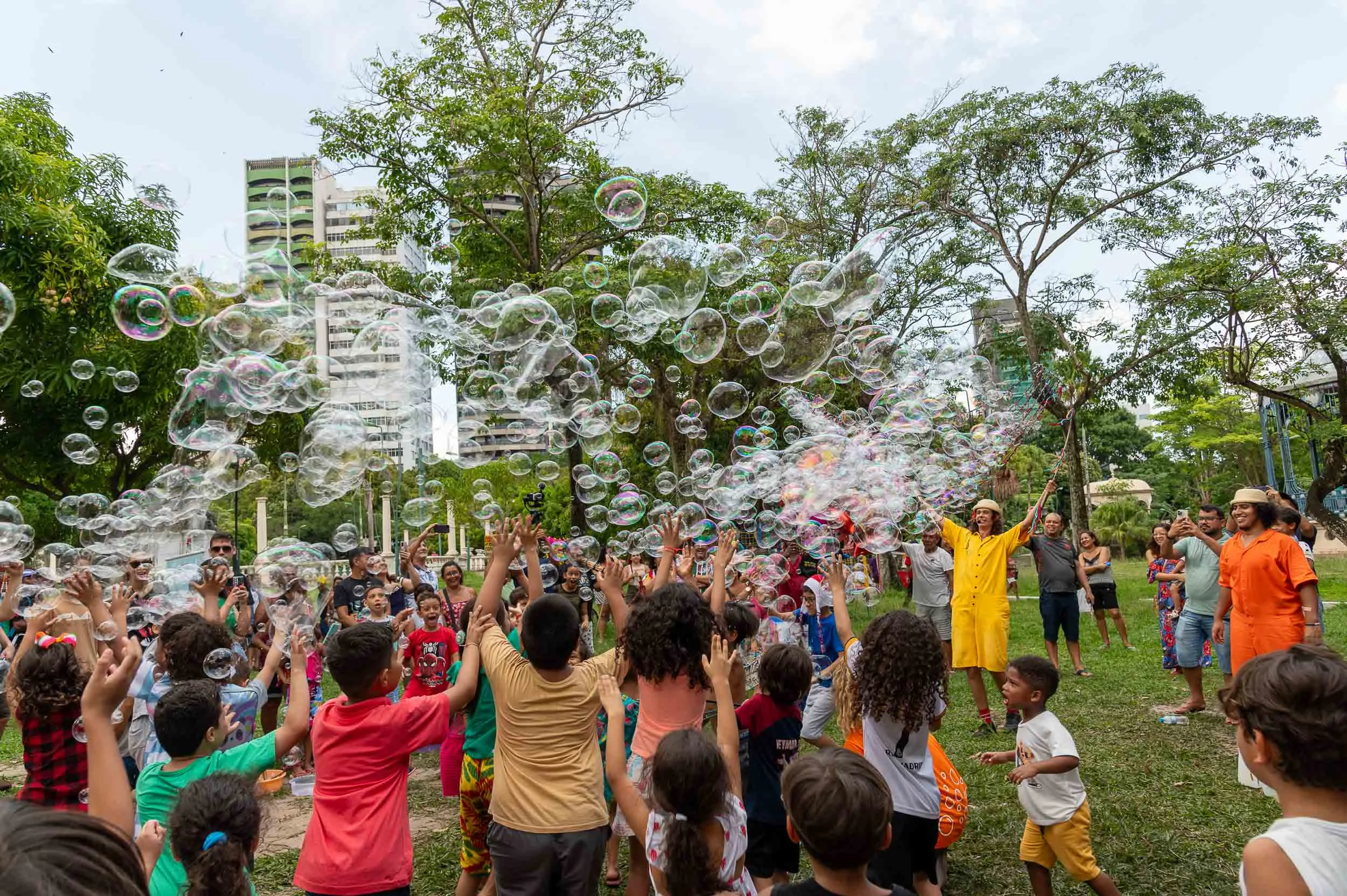 O artista Pedro Lindmayer, o Pedro Bolha, brinca com grupo de crianças fazendo bolhas de sabão ao ar livre em uma praça.